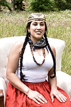 A photo of Maya Esquivido-Strom sitting outside in the grass wearing a white top and orange skirt.